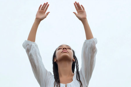 Young woman raising hands up with closed eyes, expressing spirituality, hope, prayer, and connection with nature or selfの素材