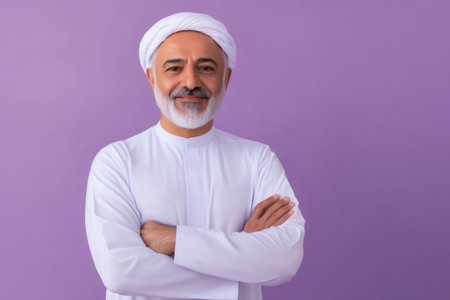 Middle Eastern man with a gray beard and traditional dress smiling confidently, standing with arms crossed against a purple backgroundの素材