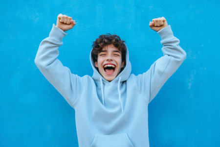 Happy teenager with a wide smile and raised fists expressing excitement and winning success against a blue backgroundの素材