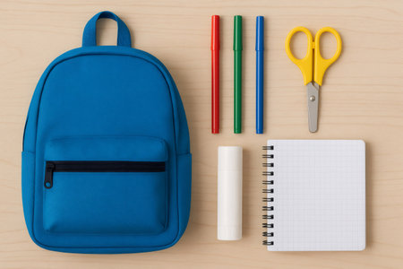 School supplies arranged on a wooden background, showing a blue backpack, colorful markers, scissors, a glue stick, and a notebookの素材