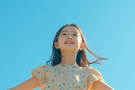 Cheerful young Asian girl smiling, looking up with hope and optimism under a bright, sunny outdoor skyの素材