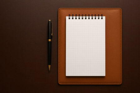 Professional overhead view of a spiral notebook with graph paper, a gold and black pen, on a brown leather desk padの素材