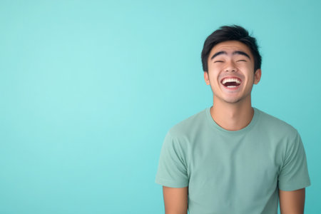 Young Asian man laughing with eyes closed against a vibrant blue background, expressing happiness and cheerful emotionの素材