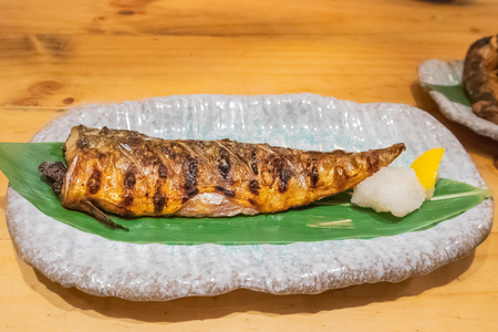 Saba grilled salt fish with green leaves on the ceramic plate on wooden table in Japanese restaurantの写真素材