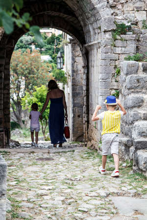 Little boy in a blue hat catches up with his mother and sister gone forward. Family walks among the ruins of the old town in Budva, Montenegroの写真素材