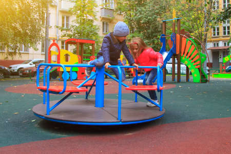 girl in pink jacket rolls boy on carousel in yard at playground. children having fun outdoors