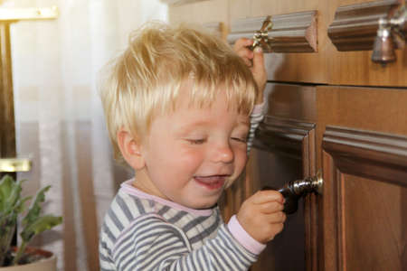 Cute little boy making first steps looked closely at handle of wooden cabinet. happy baby stands and smiles at homeの写真素材