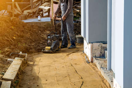 worker compresses sand in blind area around building with special working tool tamping machine or vibratory plateの写真素材