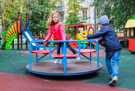 boy rolls girl in pink jacket on carousel in yard at playground outdoors