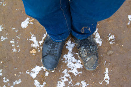 top view of black boots and legs in blue jeans all covered in mud standing on dirty ground covered...