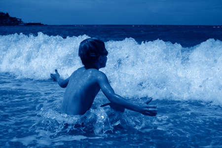 little boy in swimming goggles sitting on pebble on beach and enjoys surging waves with white foamの写真素材