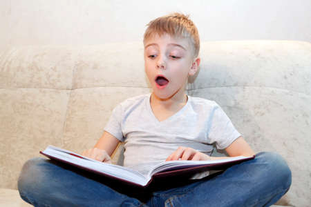 Little boy reads book sitting at home on beige sofa and is very interested in its contents