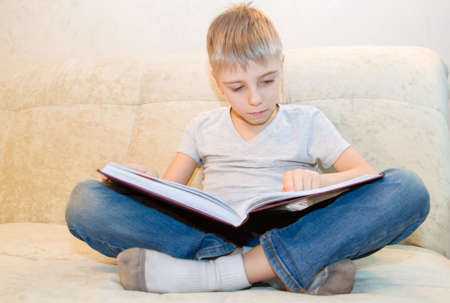 Little boy reads book sitting at home on beige sofa and is very interested in its contentsの写真素材