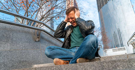 Millennial man touches his head by his hands sitting on stairs outdoors in urban area. Worried man...