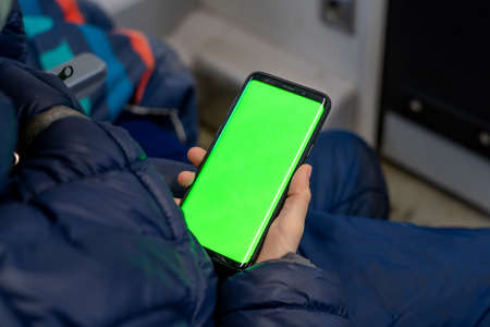 Back view of kid watch smart phone with green screen and browsing online in train. Mock up for watching content on cell phone. Blank digital phone in hand of boy stitting indoors in public transportの写真素材