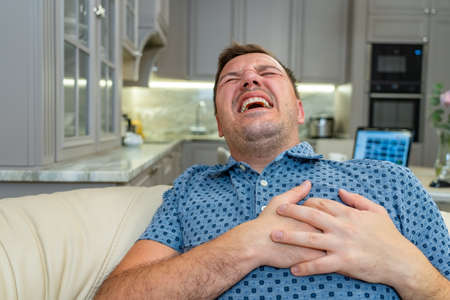 Millennial man on sofa in living room having pain in heart. Mature unshaven man suffering from heartache at remote working desk. Stressed, tired and exhausted at home officeの写真素材