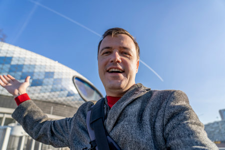 Cheerful millennial male posing to phone while making photos outside at stadium. Mature caucasian...