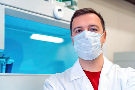 Portrait of doctor in medical mask in room with laboratory box in UV light. Tired successful male worker of scientific laboratory sitting by laboratory shelf inside office in front of cameraの写真素材