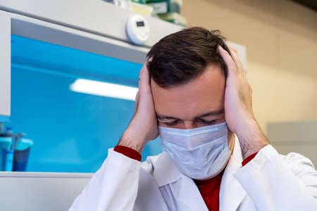 Millennial guy suffering migraine headache problem holding head with hands. Middle age professional scientist man wearing white coat with hand on head for pain in head because stress in laboratoryの写真素材