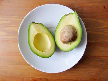 Close up fresh organic avocado halves, one with a bone, lie on a white plate on a wooden table, flat...