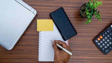 Writing on a notepad while working from home. A lap top, a mobile, calculator and plant are also on display on this brown striped working table.の写真素材
