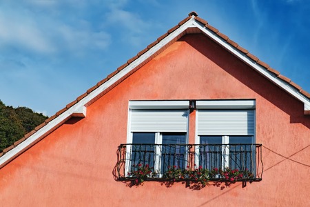 Attic windows of red stone house decorated with beautiful bright red flowers under blue sky. Gable roof and dormer type of roof outdoors.の写真素材