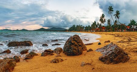 Cloudy evening view of Maenam beach, Koh Samui, Thailand. Beautiful seascape with sea waves and rocks scattered on beach sand. Rainy ominous grey storm clouds - dramatic sky.の写真素材