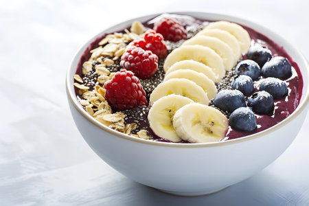 High-resolution image showcasing a colorful acai bowl garnished with ripe bananas, juicy blueberries, fresh raspberries, and a sprinkling of granola, almonds, and chia seeds, served in a speckled ceramic bowl on a white backgroundの素材