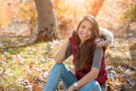 Young happy woman in park sitting among yellow leaves and smiling.