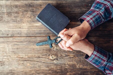 Hands are holding a crucifix and an old hard covered holly Bible and praying on a rustic wooden...