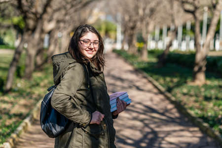 Intellectual young girl stays and look to someone in a park with some books in her hands. High...