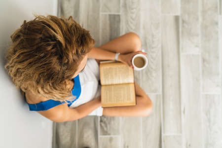 Intellectual young woman sit on floor and drink a cup of coffee, while she is reading a book. High quality photoの写真素材