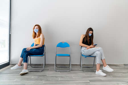 Two young woman with protection mask is sitting in a waiting hall with a distance.