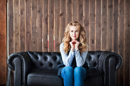 A beautiful girl in a blue shirt and a black jacket. Girl with beautiful blond curls on a black leather sofa on a wooden wall background. Business Girlの写真素材