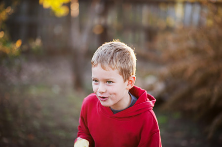 A cheerful boy with an apple rejoices, jumps and dances in summer in nature in the rays of the setting sun. A cheerful child in a red sweater and black pants. A boy eats an apple and grimacesの写真素材