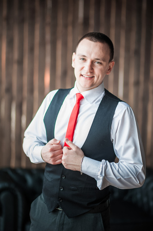 Portrait of a man in a white shirt, red tie and black vest on the background of a brick wall and a large window. Portrait of a man who is lost in thought, waiting and looking at the cameraの写真素材