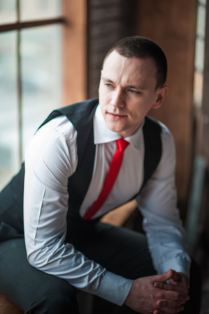 Portrait of a man in a white shirt, red tie and black vest on the background of a brick wall and a large window. Portrait of a man who is lost in thought, waiting and looking at the cameraの写真素材