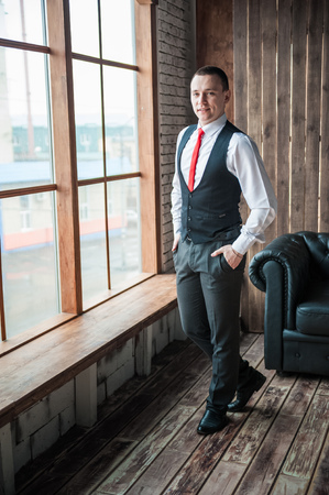 Portrait of a man in a white shirt, red tie and black vest on the background of a brick wall and a large window. Portrait of a man who is lost in thought, waiting and looking at the cameraの写真素材