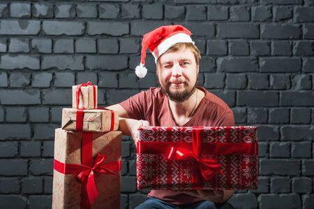 A man in a burgundy T-shirt, Santa Claus hat. A funny bearded guy with lots of gifts on different backgrounds.の写真素材