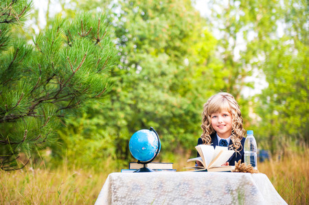 The girl is a first grader sitting at a table and holding an open book in her hands. A schoolgirl in a blue suit and curly hair on the last of the first or the first bell in the background of nature. Globe and a bottlの写真素材