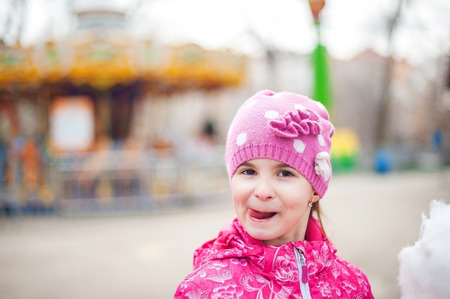 A photograph of a child's rest on nature in the spring. A child in a bright pink jacket is wearing a sweet cotton wool against the backdrop of rides, carousels. Close-up portrait with sweet cotton wの写真素材