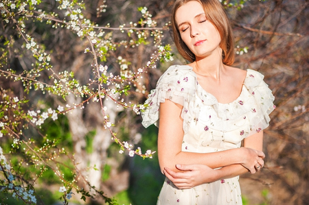 Beautiful slim girl with fair-haired hair in a long light dress against the backdrop of nature, flowering trees, at home, in the summer, in the springの写真素材