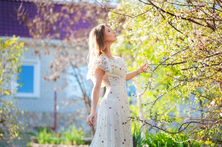 Beautiful slim girl with fair-haired hair in a long light dress against the backdrop of nature, flowering trees, at home, in the summer, in the springの写真素材
