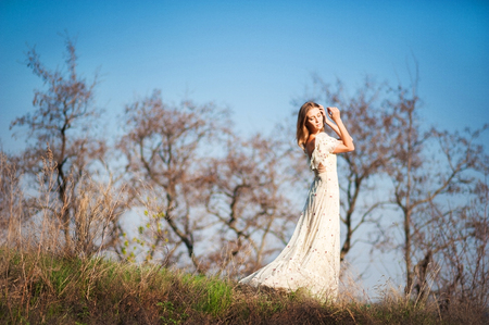 Beautiful slender girl with light brown hair in a long light dress on a background of nature, dry grass, green grass, blue sky in summer, in the springの写真素材