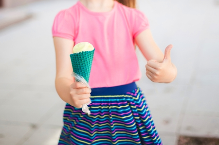 A girl in a pink T-shirt and a bright multi-colored skirt is holding ice cream in a green horn. Ice...