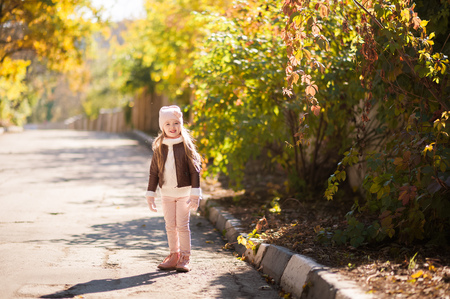 Autumn children's fashion. A little girl dances, jumps and rejoices in the fall against a background of yellow and red foliage on trees and bushes. Joy and happiness in childhoodの写真素材