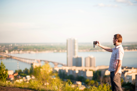 A man in a shirt and trousers takes pictures of himself and nature on the phone. Portrait of a man in the background of Saratov, Russia, view of the houses, the Volga River, the bridge to Engels.の写真素材