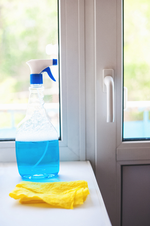 A blue window cleaner, a yellow rag on a white window. Carrying out home inspections. Dirty and clean window, detergent, the concept of houseworkの写真素材