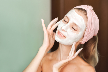 Beauty Treatments. Young woman is applying a mask, cream on her face closeup. Facial skin care portrait of a beautiful girl with a towel on her head applying facial maskの写真素材