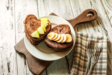 Heart-shaped chocolate pancakes on a dark background. Pancake with banana, jam, apple, forks. Breakfast for your beloved on Valentine's Dayの写真素材